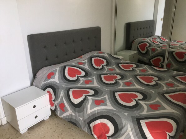 A bedroom with a grey tufted headboard, a white bedside table with drawers, and a bed with a patterned bedspread featuring heart shapes.