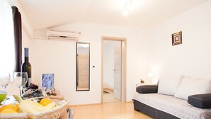 A modern hotel room with a wooden floor, a bed with a grey cover, a small table with a basket of fruit, a wine bottle, and a framed picture on the wall. - Rooms Garden Zagreb Airport (Velika Gorica)