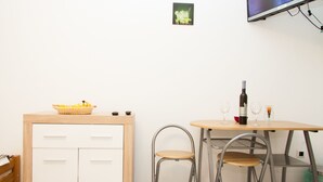 A small dining area with a wooden table, two metal chairs, a basket of fruit, a bottle of wine, and a television mounted on the wall. - Rooms Garden Zagreb Airport (Velika Gorica)
