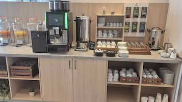 A café counter with a coffee machine, juice dispensers, and a variety of cups and mugs displayed on shelves.