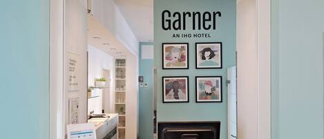 A reception area with a sign reading 'RECEPTION' and 'Garner AN IHG HOTEL'. There are framed artworks on the wall and a reception desk with a bell and a glass.