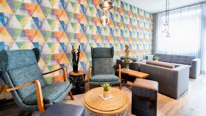 Un salón moderno con una pared de color con estampado geométrico, un sillón azul, un sofá gris y una mesa de centro redonda de madera.