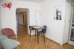 Una habitación con piso de madera, una mesa con una revista, una silla roja, una silla azul y un aplique de pared.