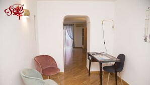 A room with a wooden floor, a table with chairs, a wall-mounted lamp, and a door leading to another room.