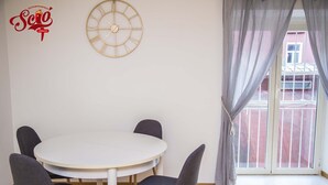 A dining area with a round table and four chairs, a wall clock, and a window with curtains.