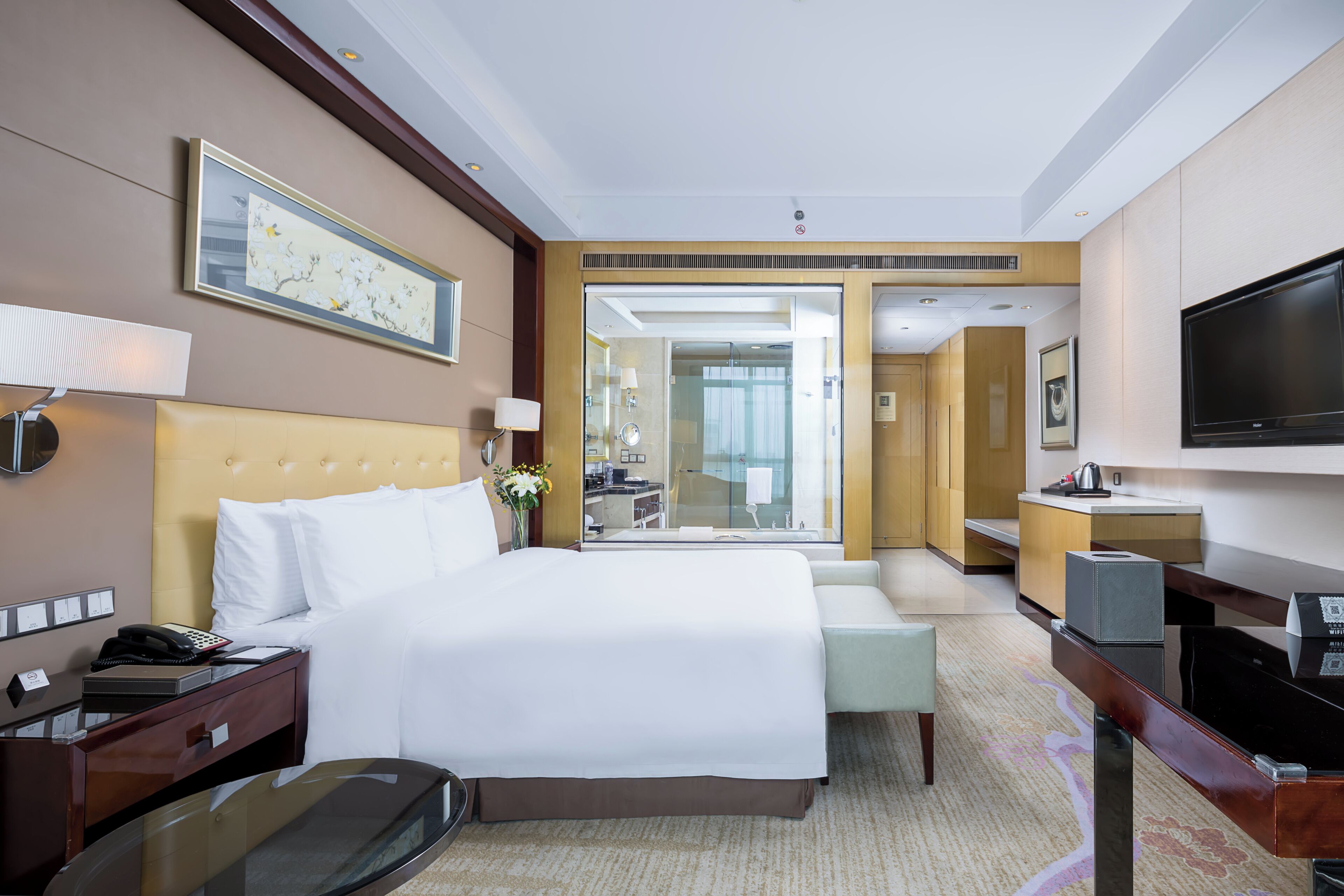 A hotel room with a large bed, bedside table, lamp, TV, and a view of the bathroom and kitchen area.