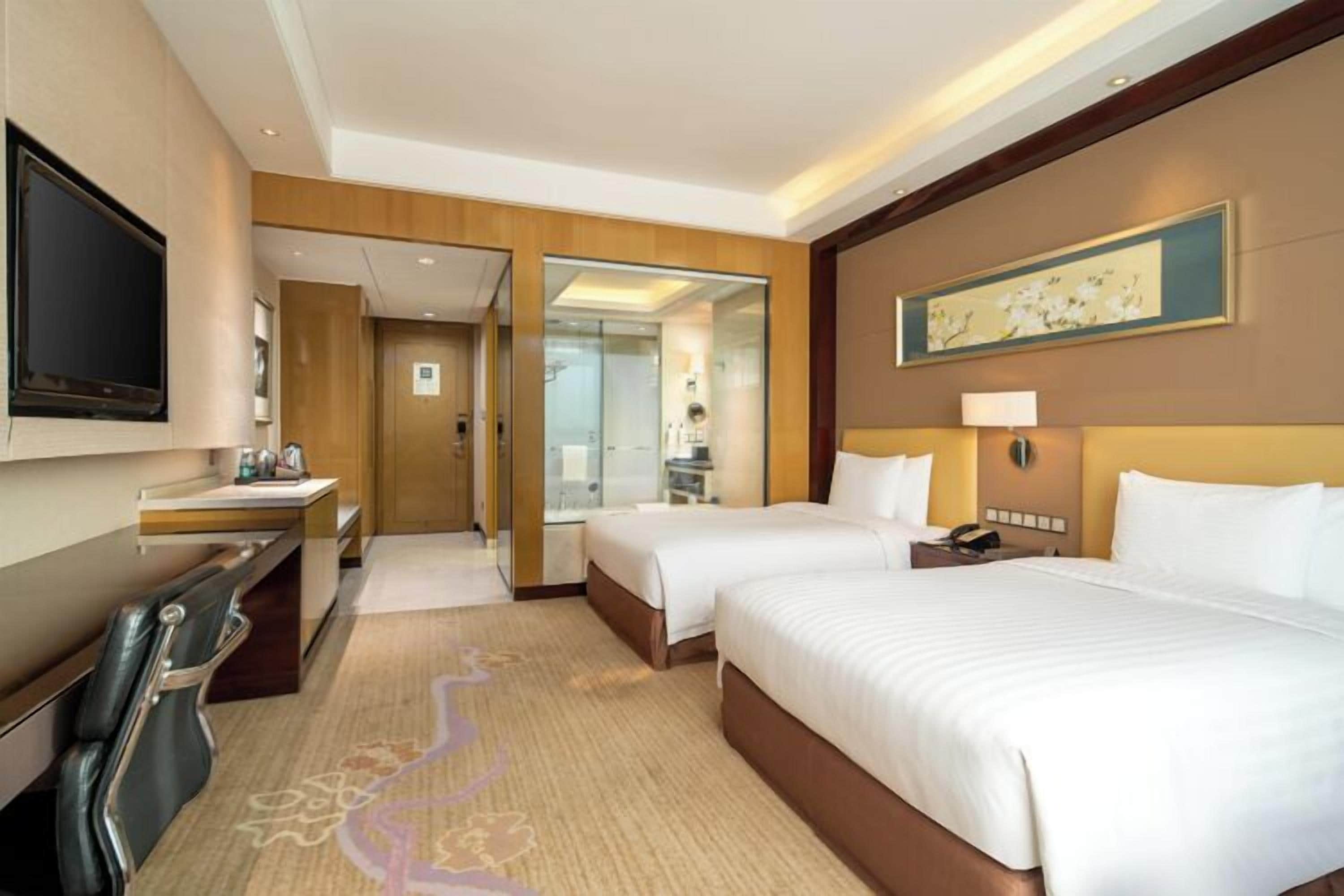 A hotel room with two beds, a desk with a chair, a TV, and a bathroom visible through an open door.