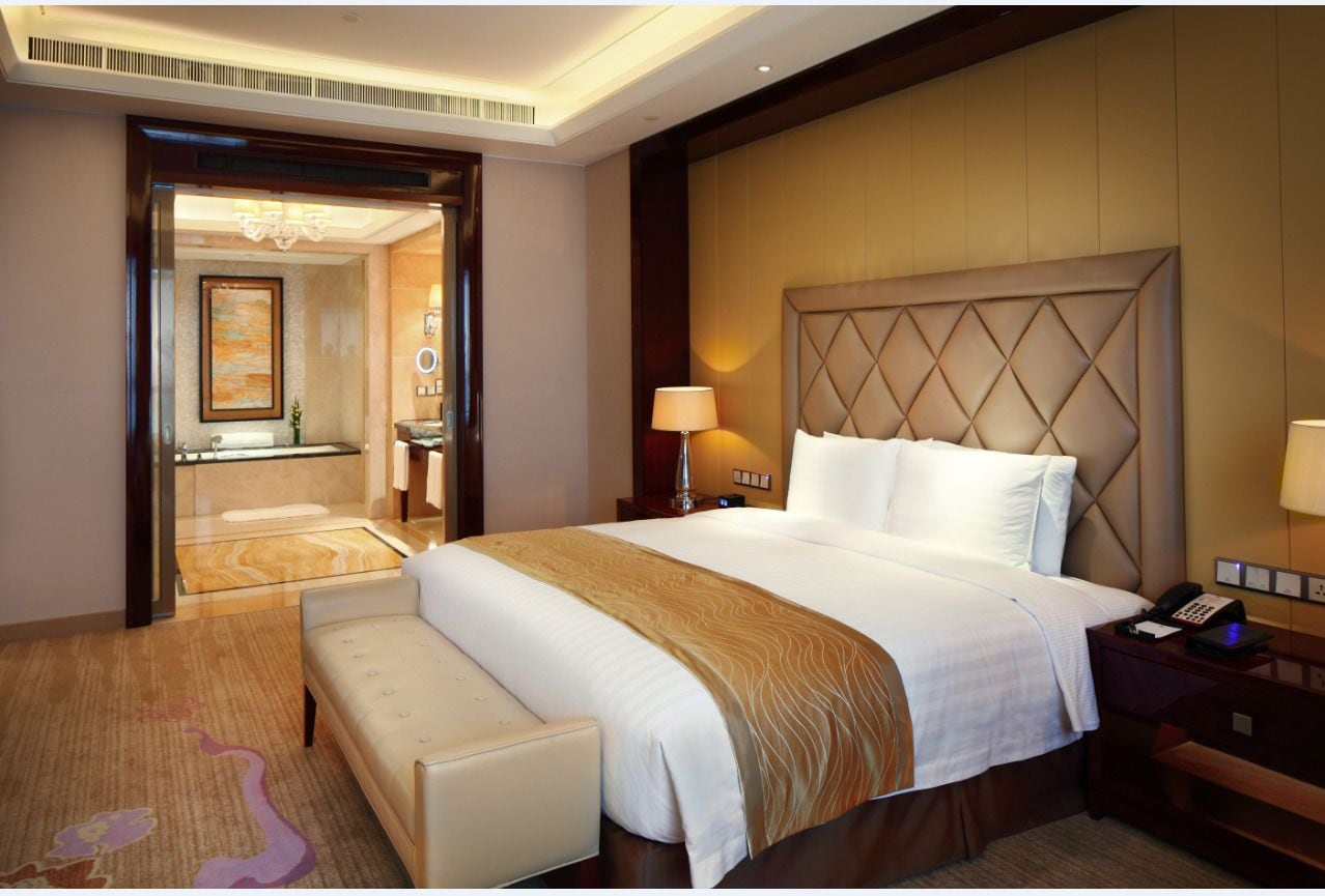 Habitación de hotel con una cama grande, una mesita de noche con lámpara, un banco y vista al baño por una puerta abierta.