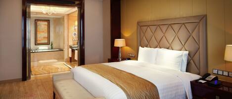 Habitación de hotel con una cama grande, una mesita de noche con lámpara, un banco y vista al baño por una puerta abierta.