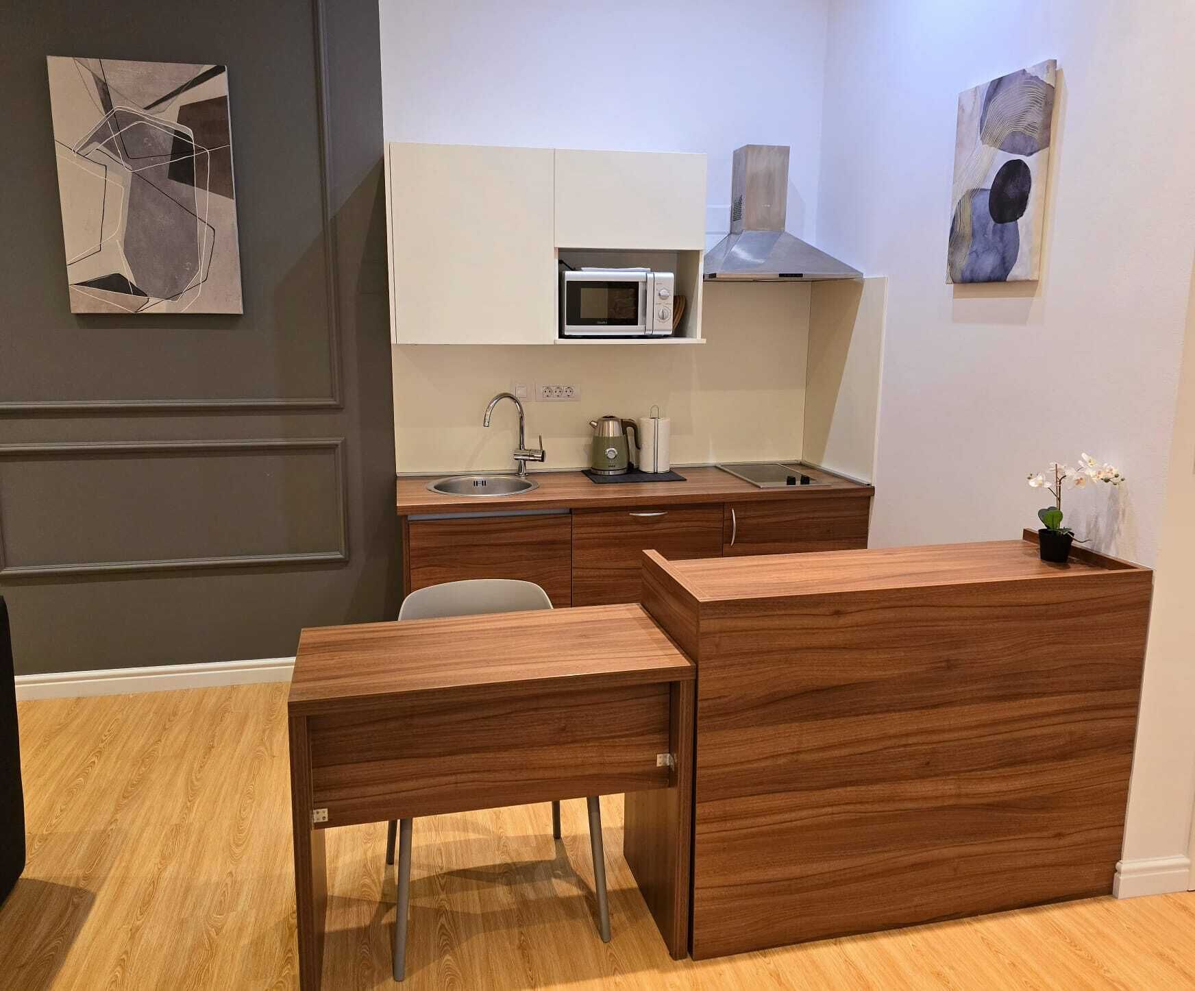 A compact kitchen with a microwave, sink, and a small countertop. There is a wooden table and a chair in the foreground.