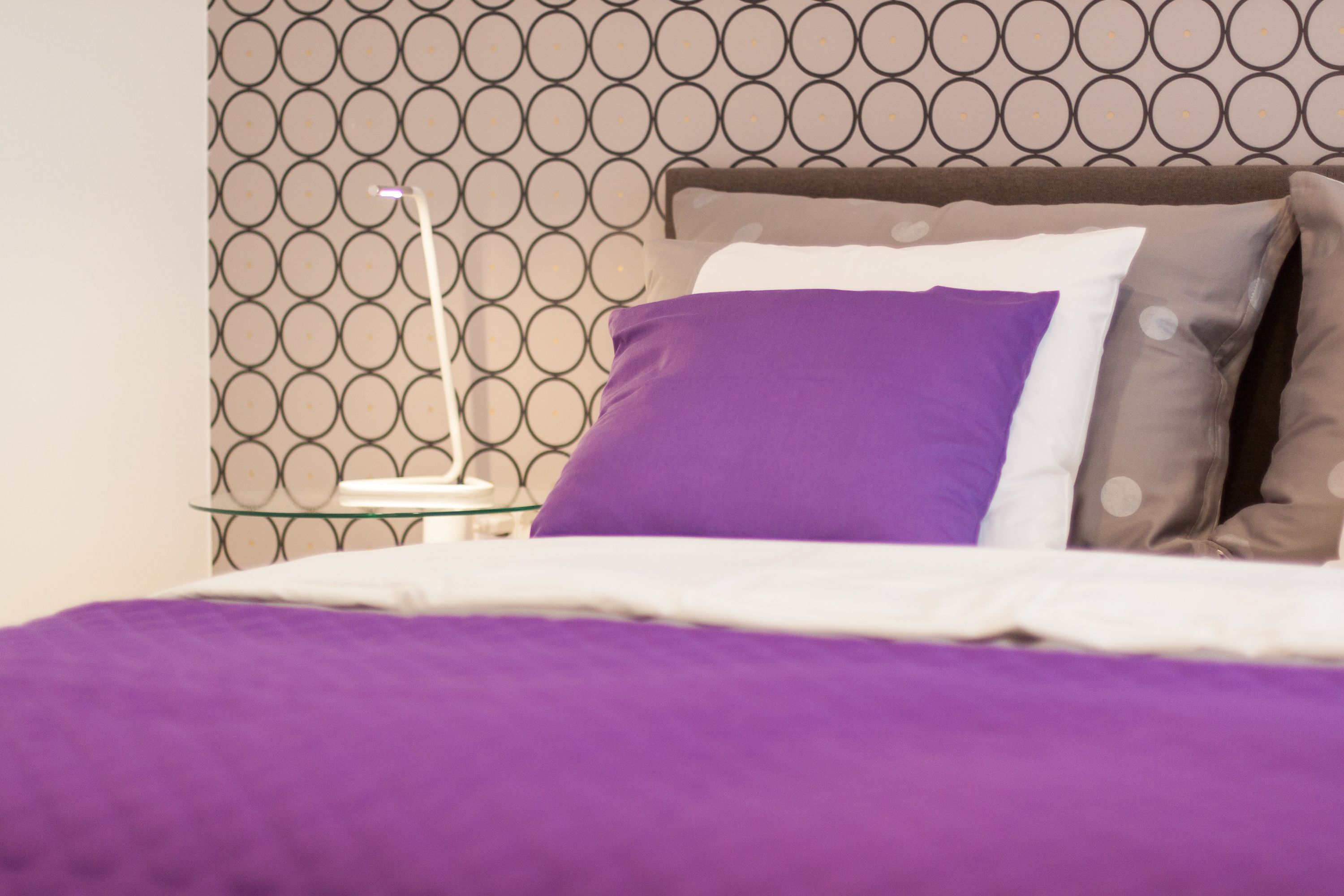 A bed with a patterned headboard, a bedside table with a lamp, and a purple blanket.