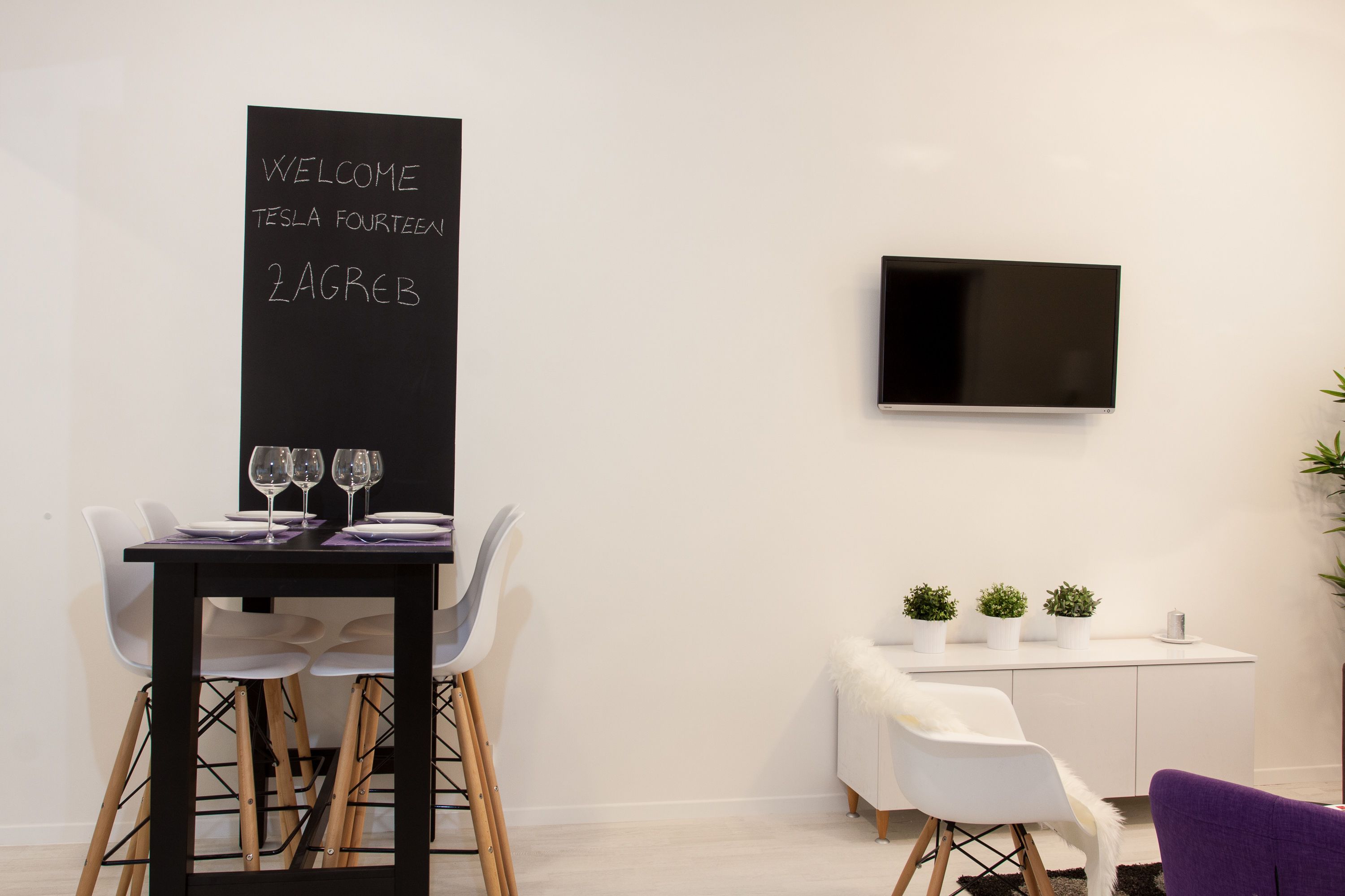 A modern room with a blackboard displaying "WELCOME TESLA FOURTEEN 2AGREB", a TV mounted on the wall, a dining table with chairs, and a small seating area with plants.