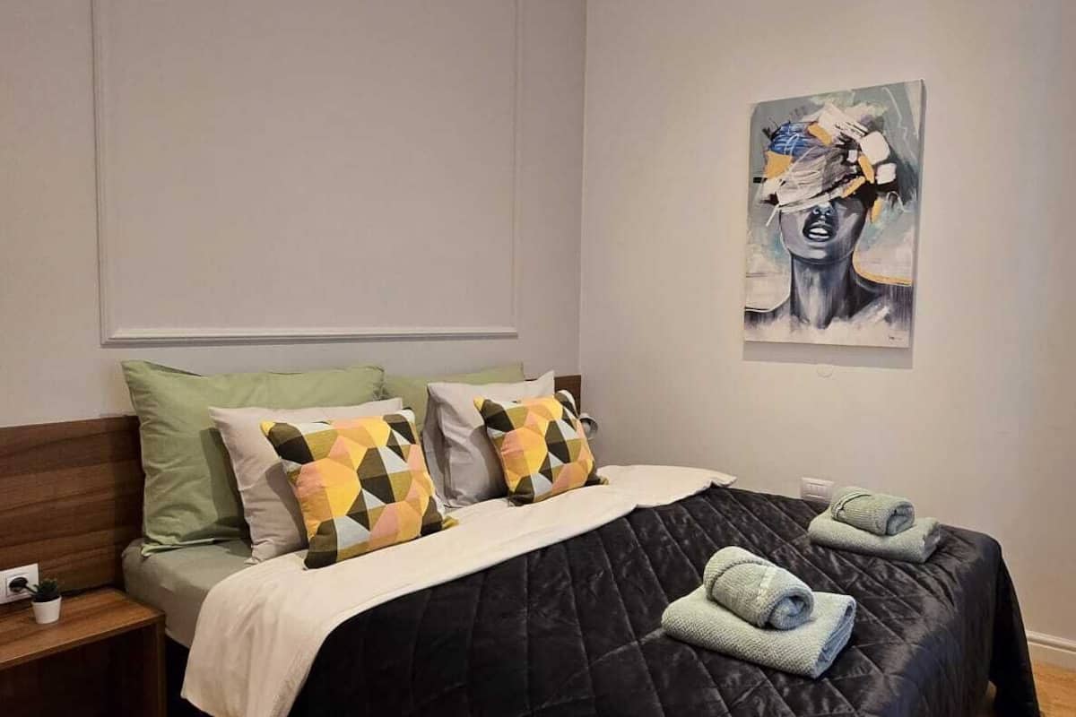 Une chambre à coucher avec une couvre-lit matelassé de couleur sombre, plusieurs oreillers, une table de chevet et un tableau accroché au mur.