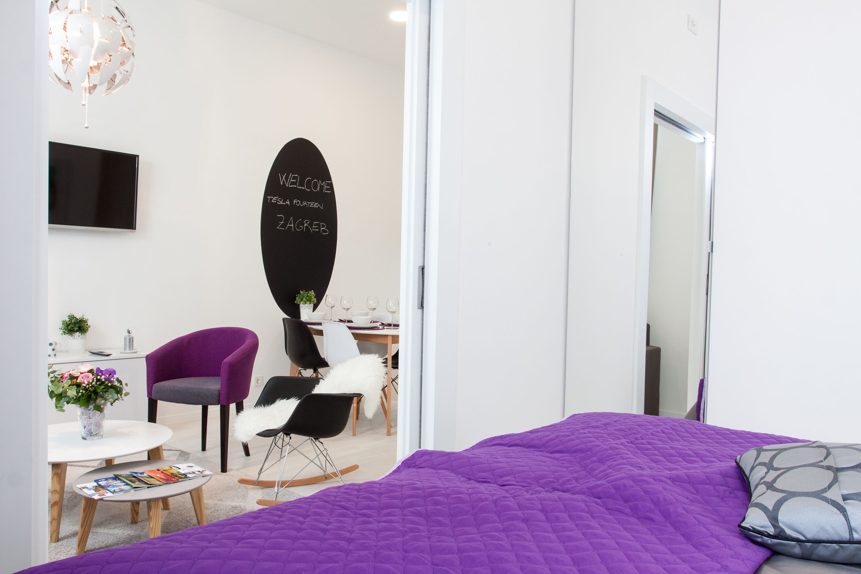 A modern hotel room with a purple bedspread, a blackboard with a welcome message, a flat-screen TV, and a dining area with a table and chairs.