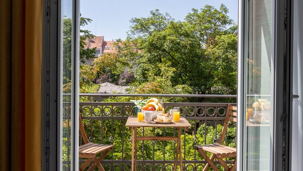 Een balkon met een ontbijttafel, compleet met fruit en sap, met uitzicht op een weelderige tuin en gebouwen.