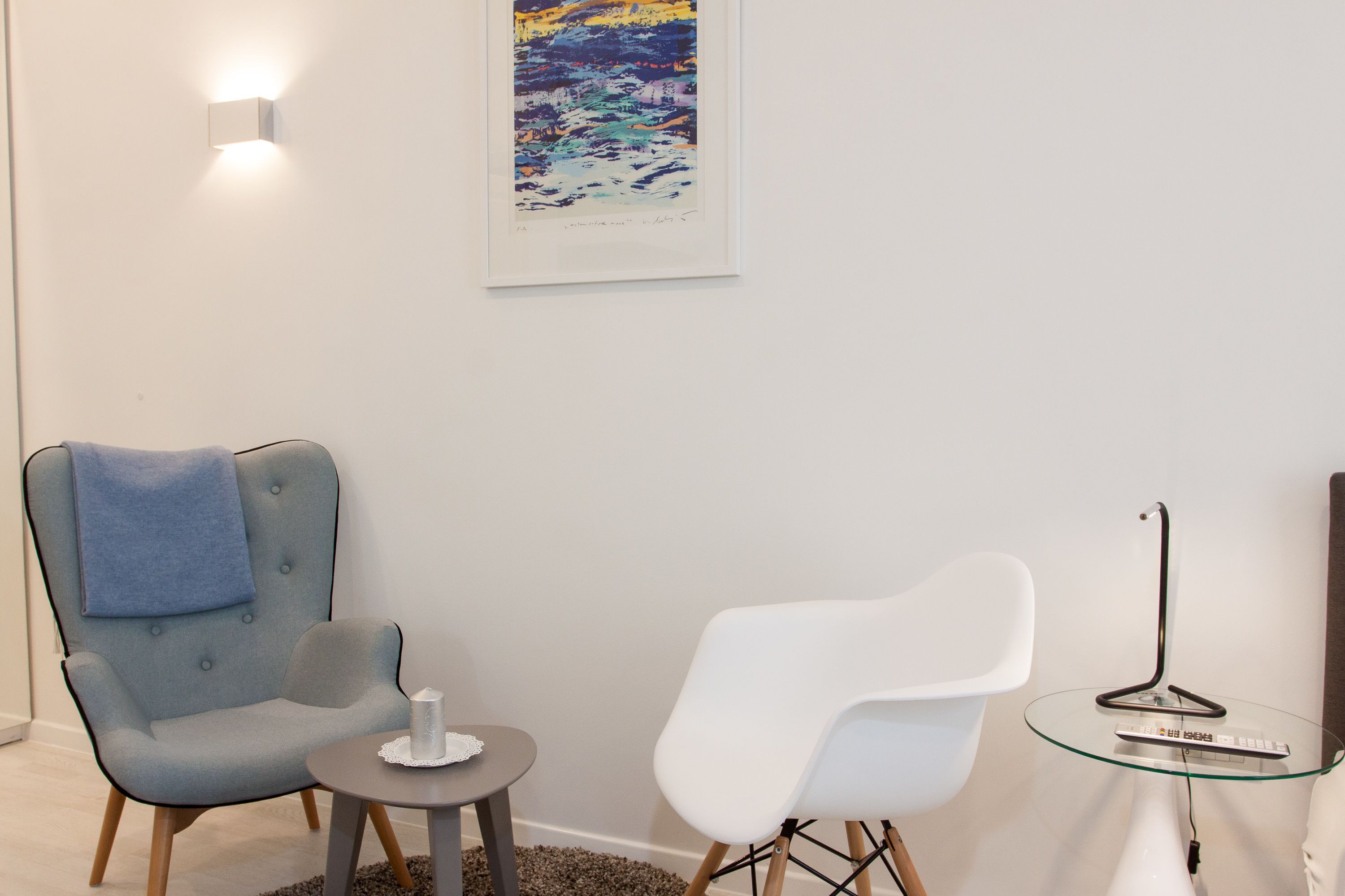 A room with a blue armchair, a white chair, a small table with a candle, and a glass table with a remote control.