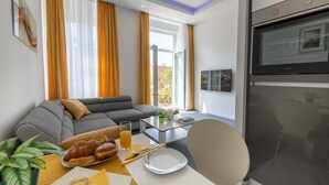 Een moderne woonkamer met een grijze bank, een eettafel met ontbijtspullen, een televisie en een groot raam met gordijnen.