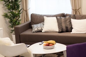Een woonkamer met een bruine bank, witte kussens, een klein wit tafeltje met een fruitschaal, en een kamerplant.