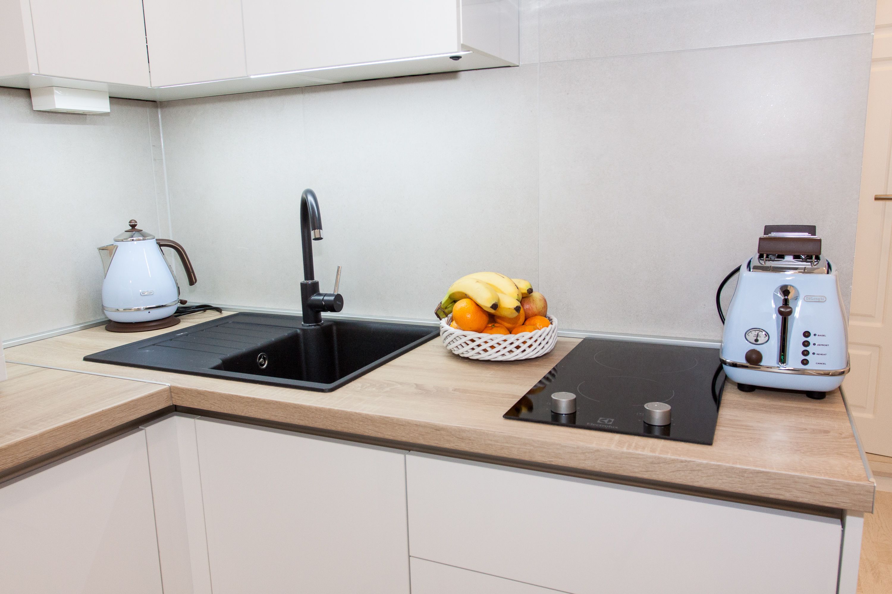 A modern kitchen with a black sink, a white kettle, a toaster, and a fruit basket on the countertop.