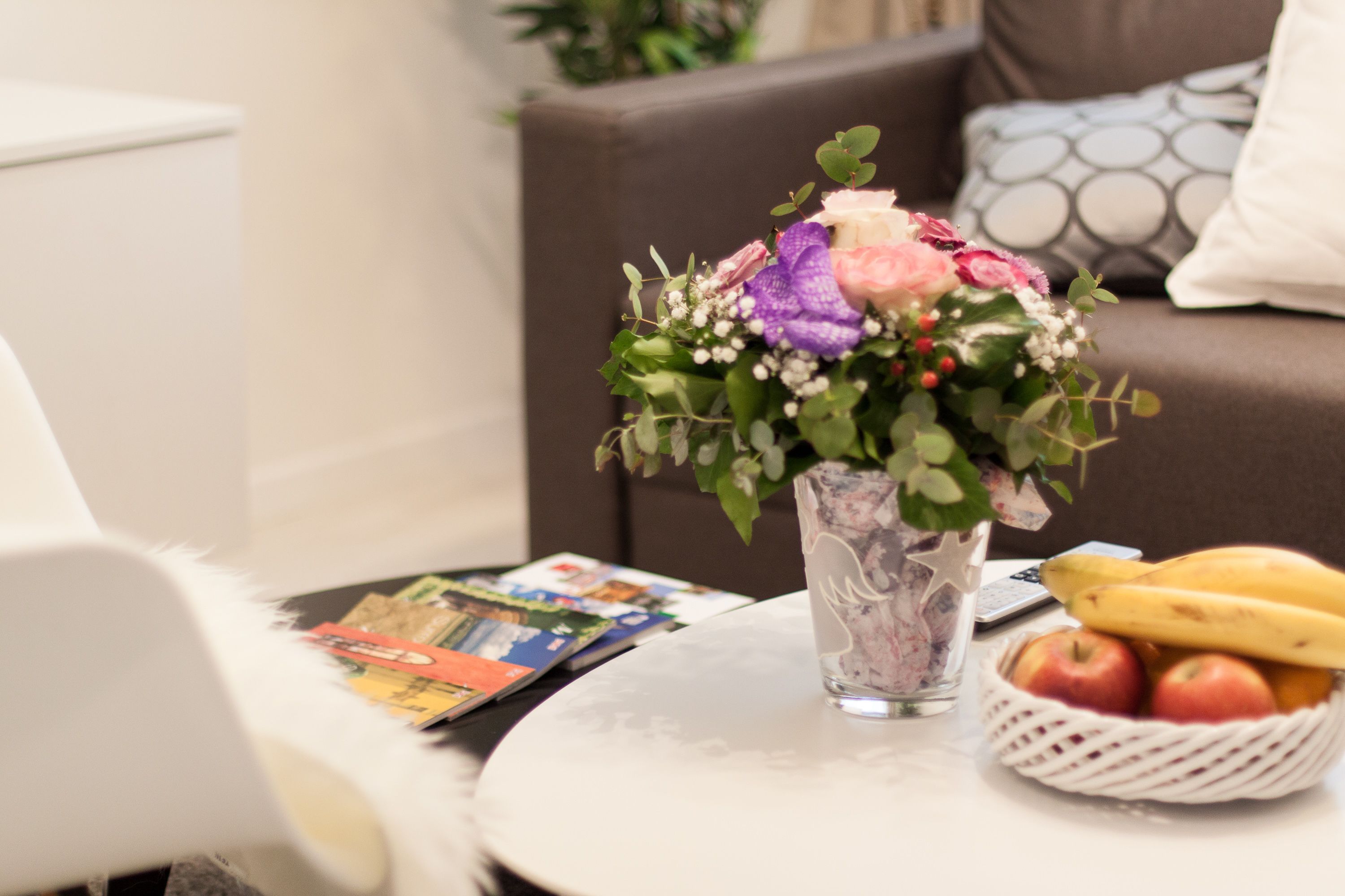 A round table with a vase of flowers, a basket of fruit, and magazines.