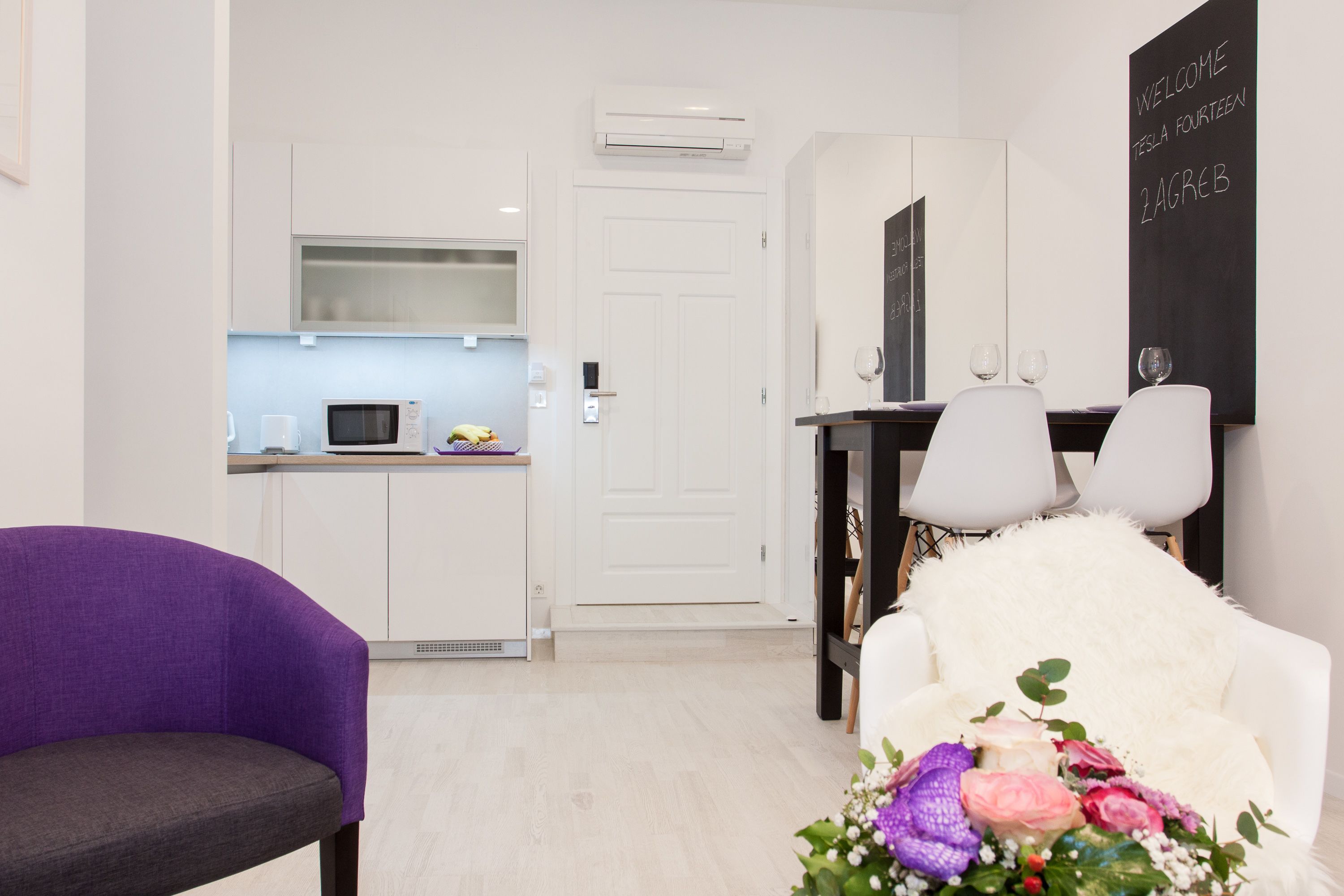 A modern hotel room with a kitchen area, a dining table with chairs, and a purple armchair.