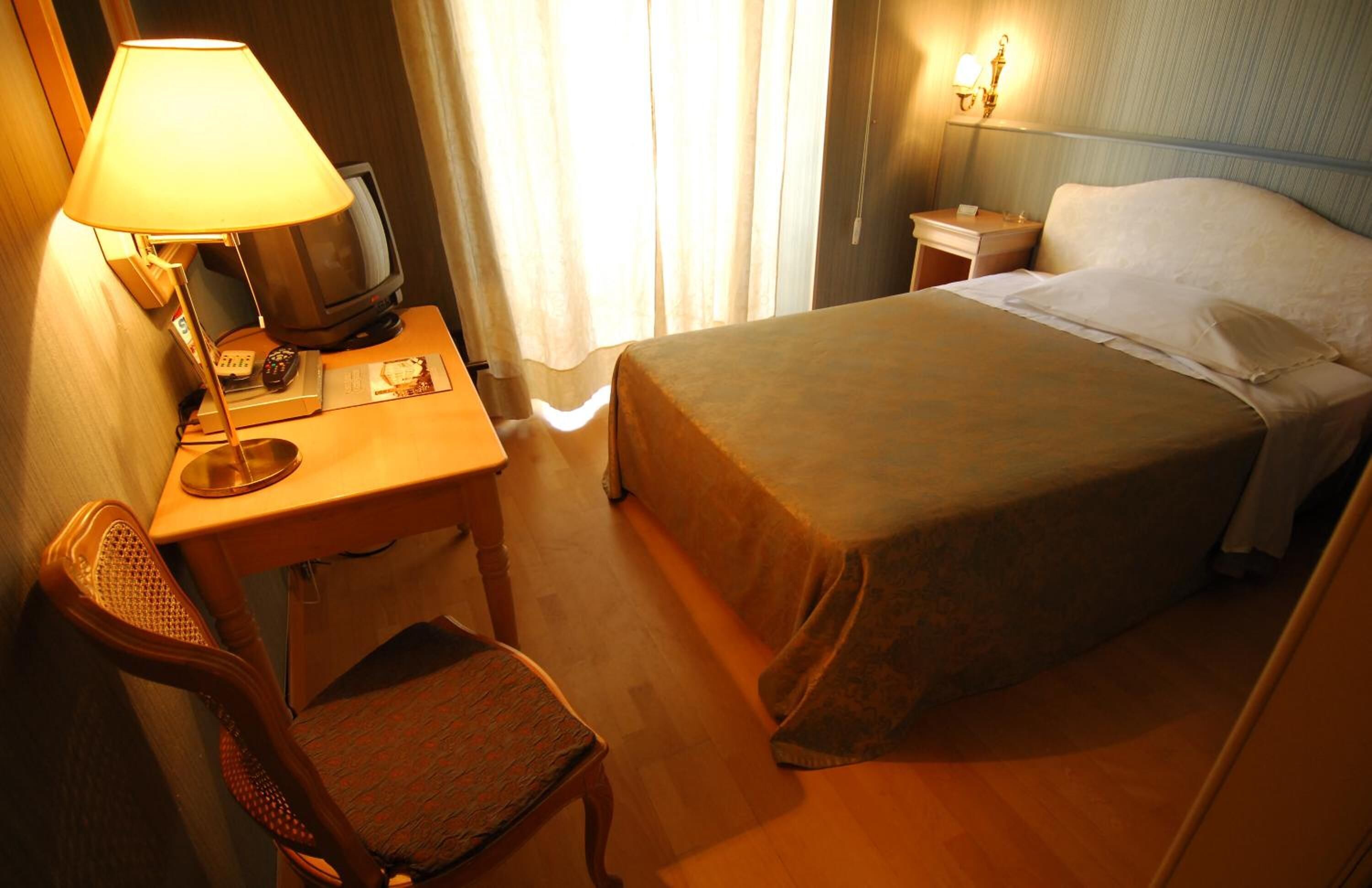 A hotel room with a bed, a desk with a lamp, a chair, a television, and a nightstand with a lamp.