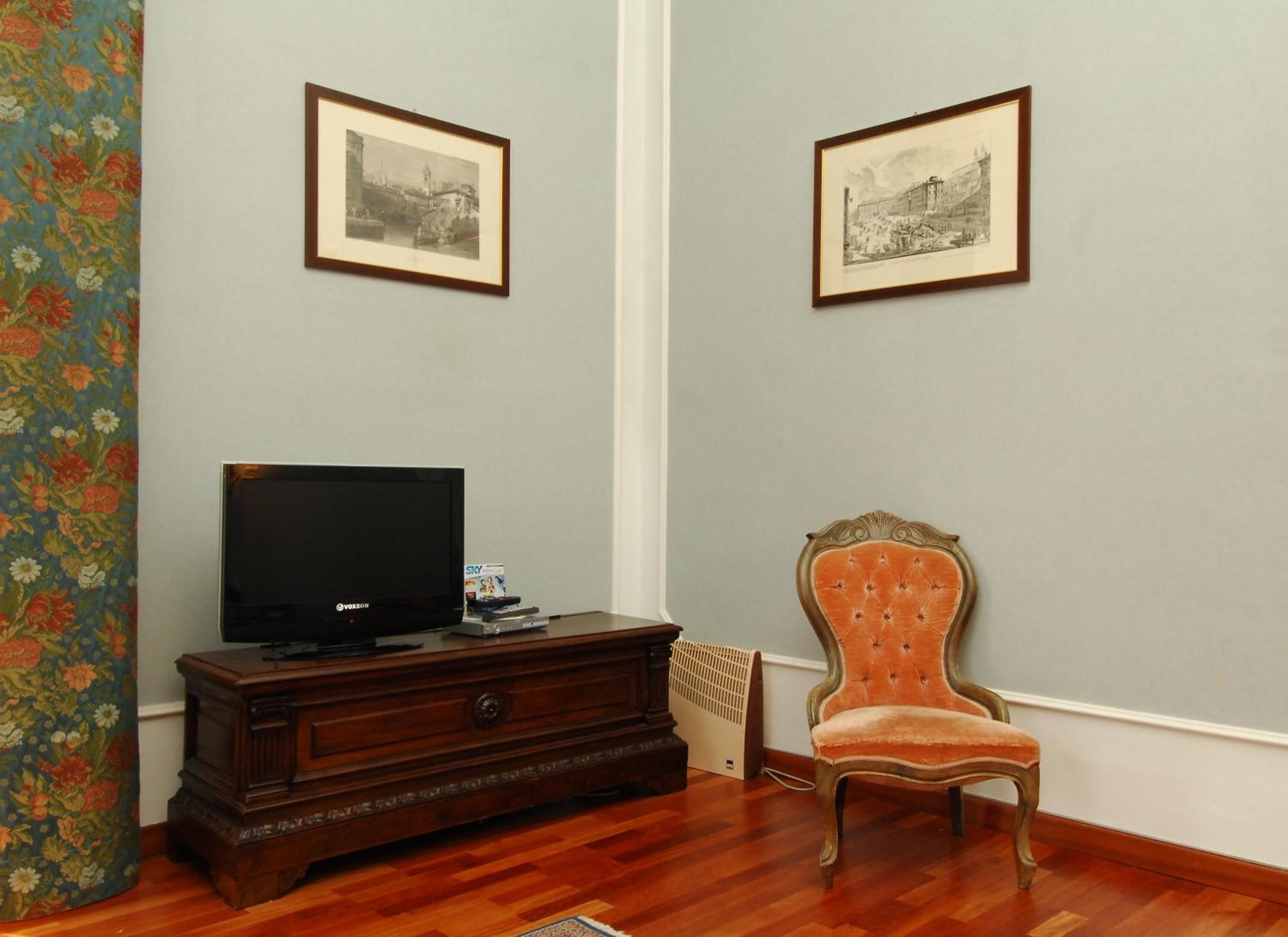 Quarto com televisão sobre um armário de madeira, uma cadeira antiga e duas fotografias a preto e branco emolduradas na parede.
