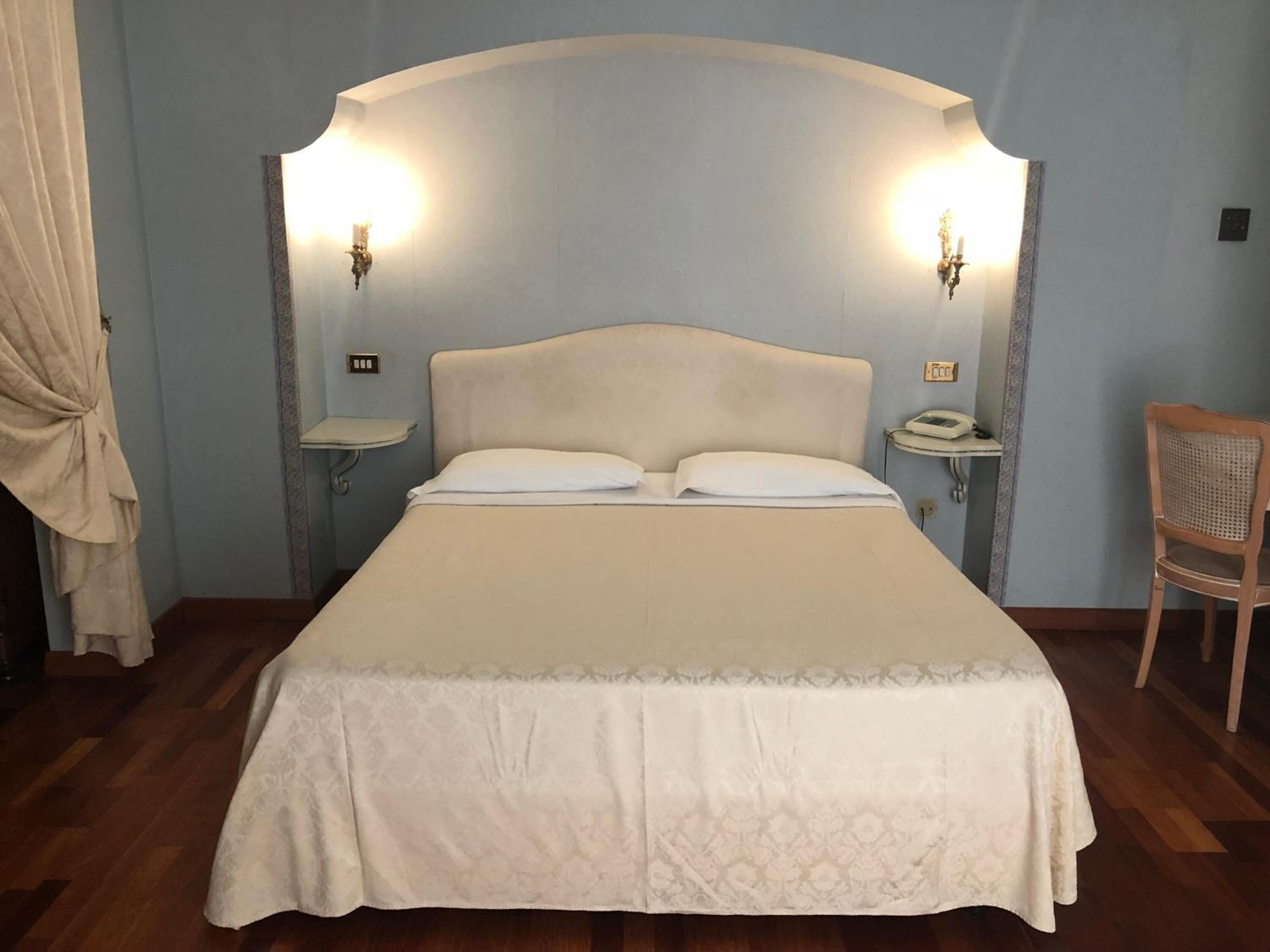 A neatly made bed with a headboard, two bedside lamps, a bedside telephone, and a wooden chair.