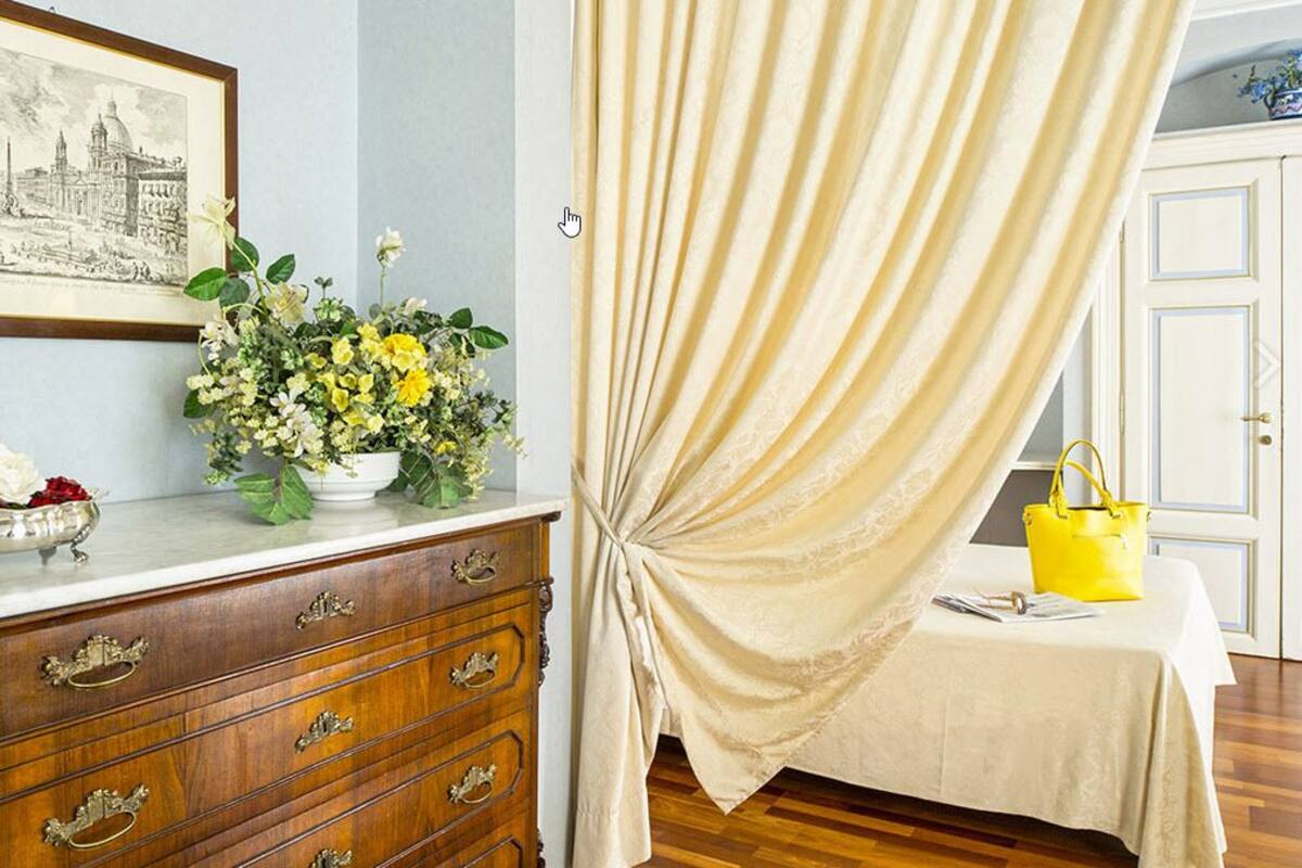A room with a wooden dresser, a framed cityscape picture, a vase of flowers, a bed with a white bedspread, and a yellow handbag on the bed.