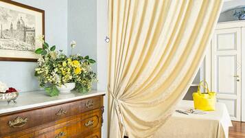 A room with a wooden dresser, a framed cityscape picture, a vase of flowers, a bed with a white bedspread, and a yellow handbag on the bed.