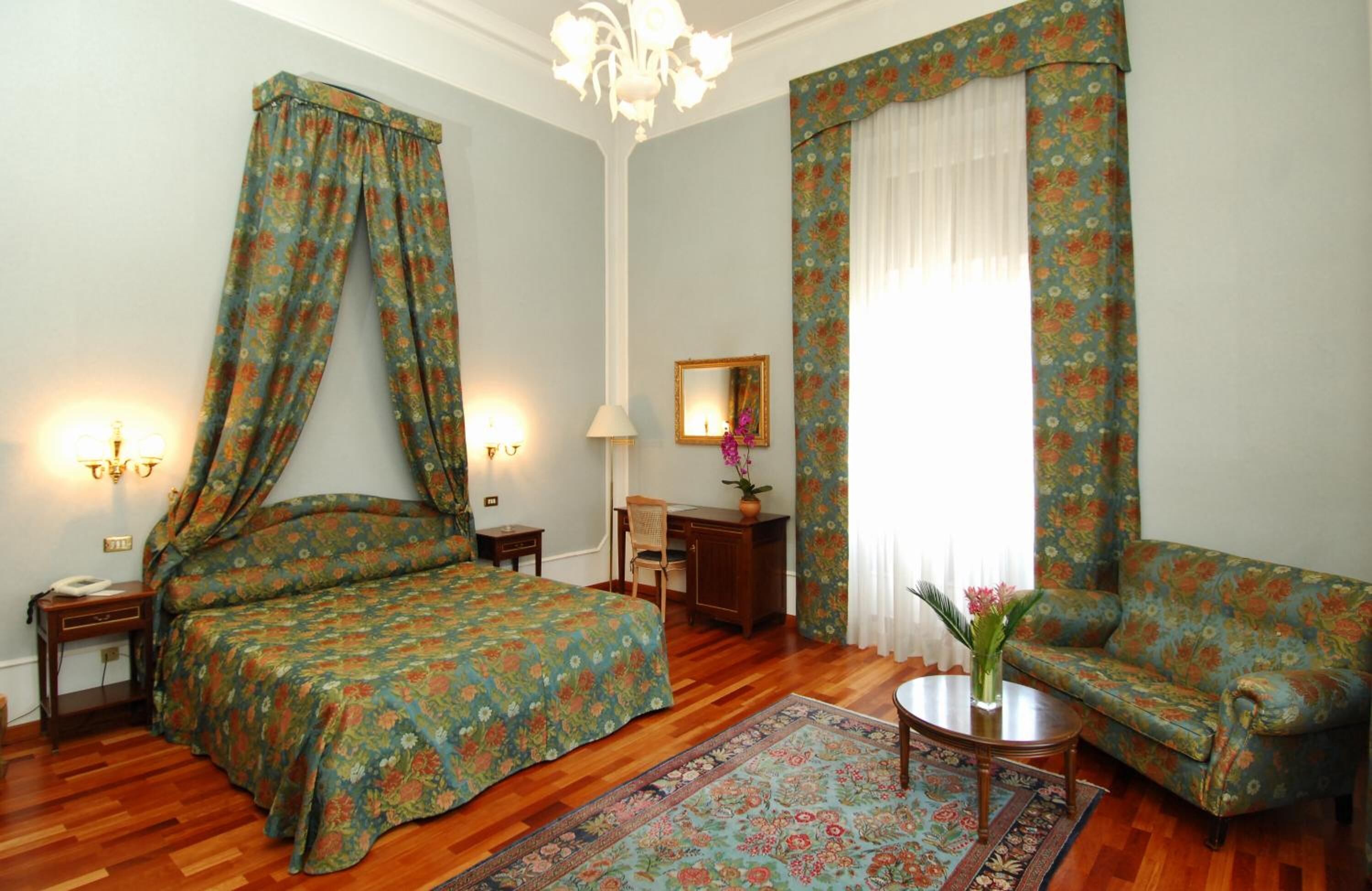 Ein Schlafzimmer mit Himmelbett, einer Sitzecke mit Stuhl und Tisch, einem Schreibtisch mit Lampe und einem großen Fenster mit Vorhängen.