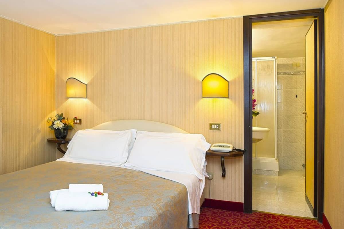 A hotel room with a bed, two wall-mounted lamps, a bedside table with a phone, and a bathroom visible through an open door.