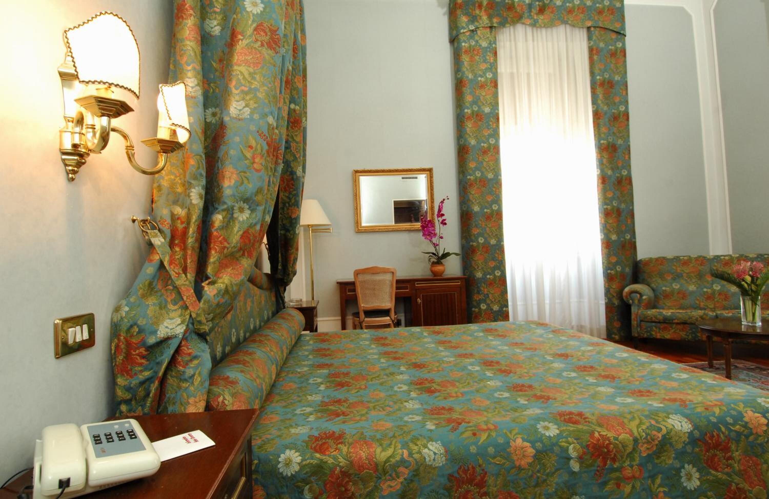 Una habitació d'hotel amb un llençol floral, un escriptori amb una làmpada, una cadira, una taula petita amb flors i un telèfon.