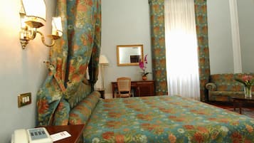 Una habitació d'hotel amb un llençol floral, un escriptori amb una làmpada, una cadira, una taula petita amb flors i un telèfon.