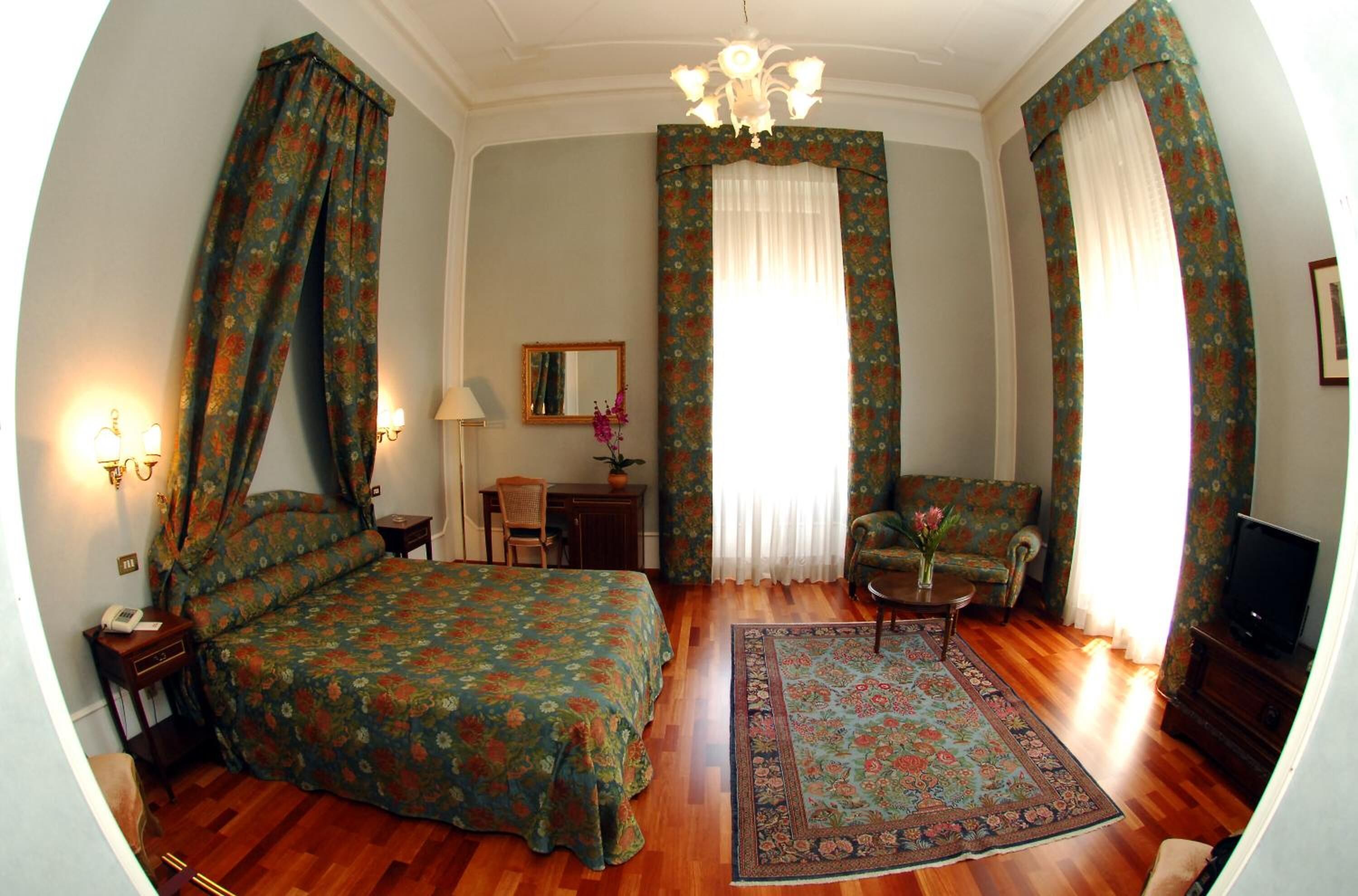A bedroom with a bed, a desk, a chair, a television, and a chandelier.