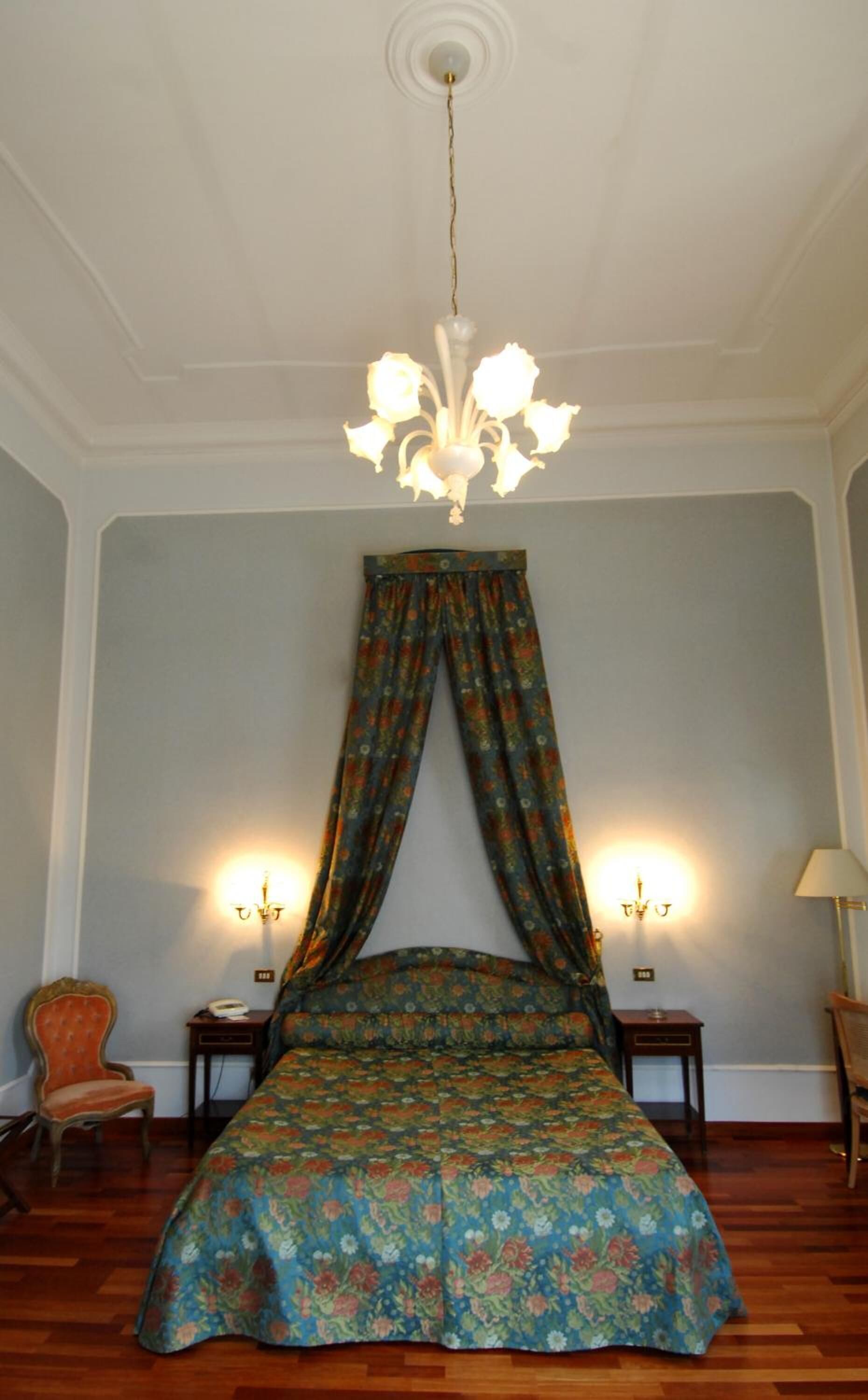 Ein Schlafzimmer mit Himmelsbett, zwei Nachttischen mit Lampen, einem Sessel und einer Kronleuchter.