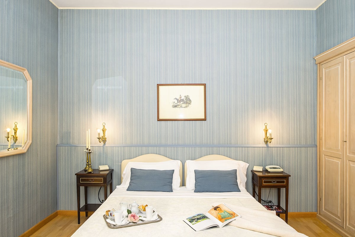 A hotel room with two beds, a bedside table with a lamp, a framed picture on the wall, and a tray with items on the bed.