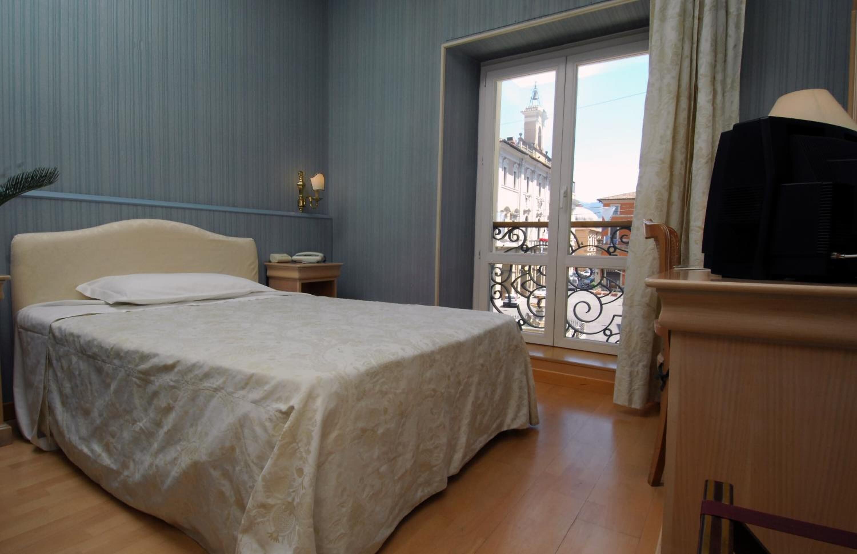 Un dormitorio con cama, televisor, mesita de noche y ventana con vistas a edificios.