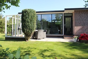Una casa moderna con un invernadero de vidrio, un patio con mobiliario exterior y un césped bien cuidado.