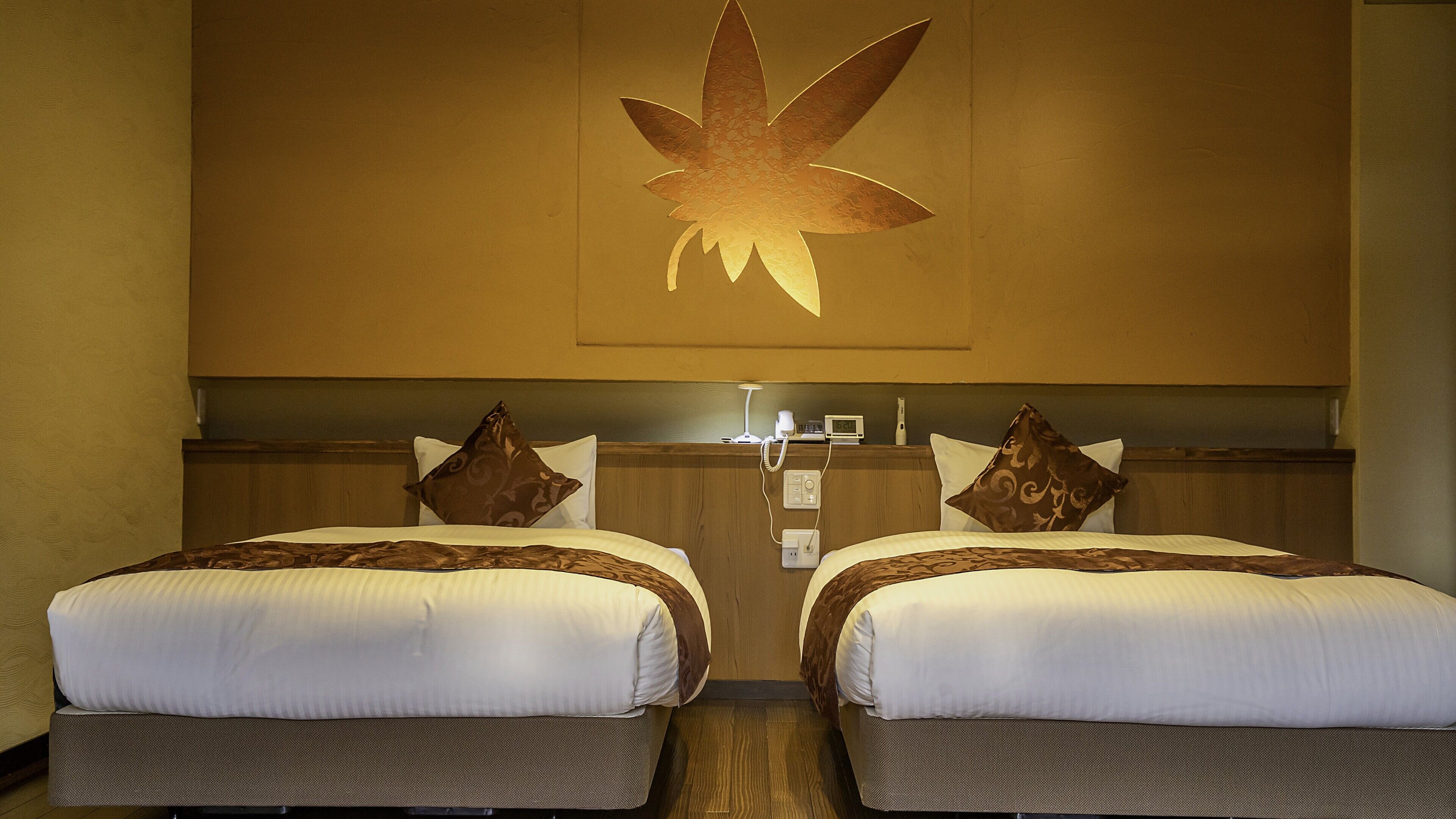 Habitación de hotel con dos camas, cada una con colcha y almohadas estampadas. Un cuadro cuelga en la pared sobre las camas, y hay una mesita de noche con una lámpara y un teléfono.
