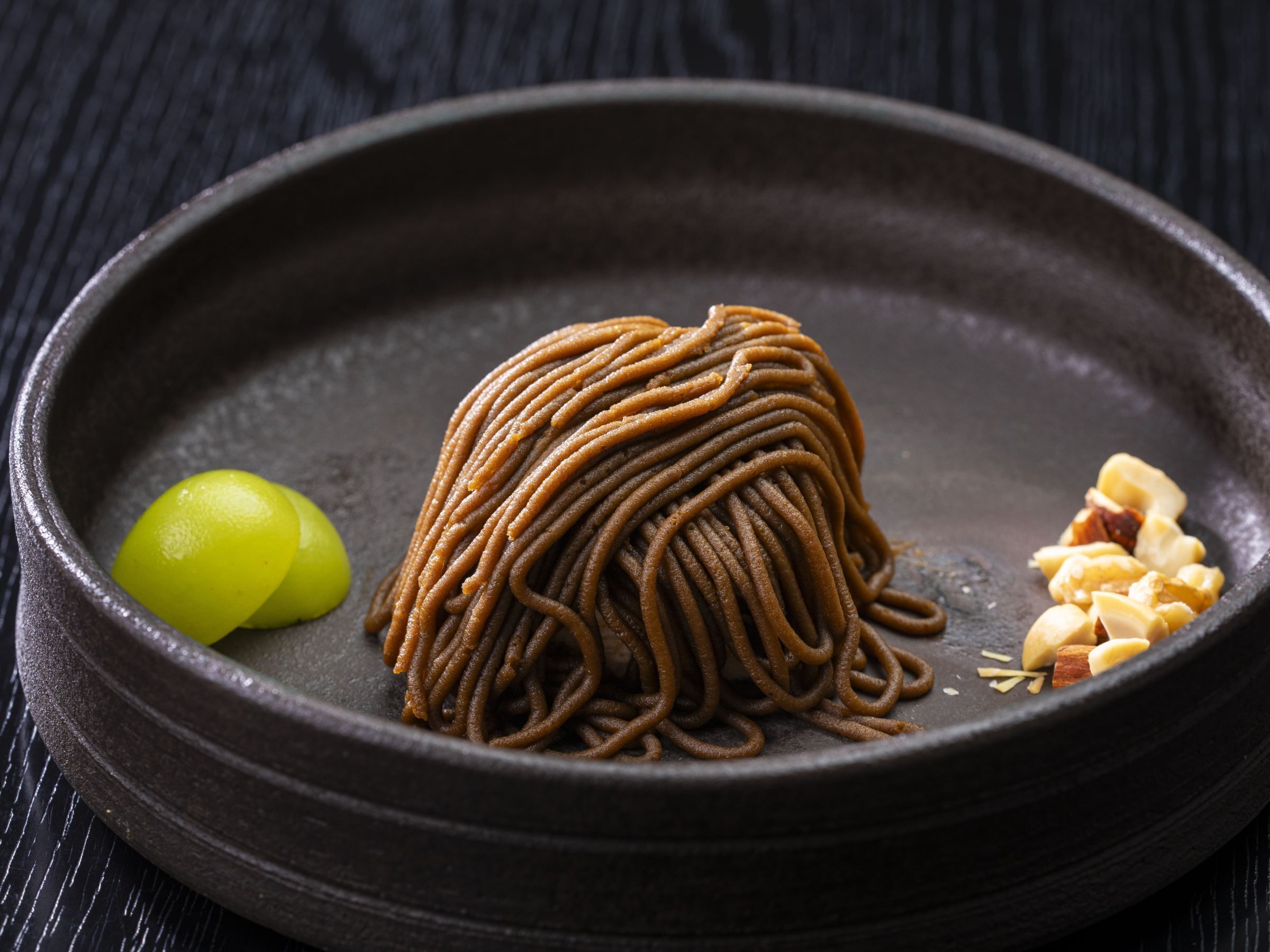 A bowl of noodles with a small portion of green grapes and peanuts on a dark plate.