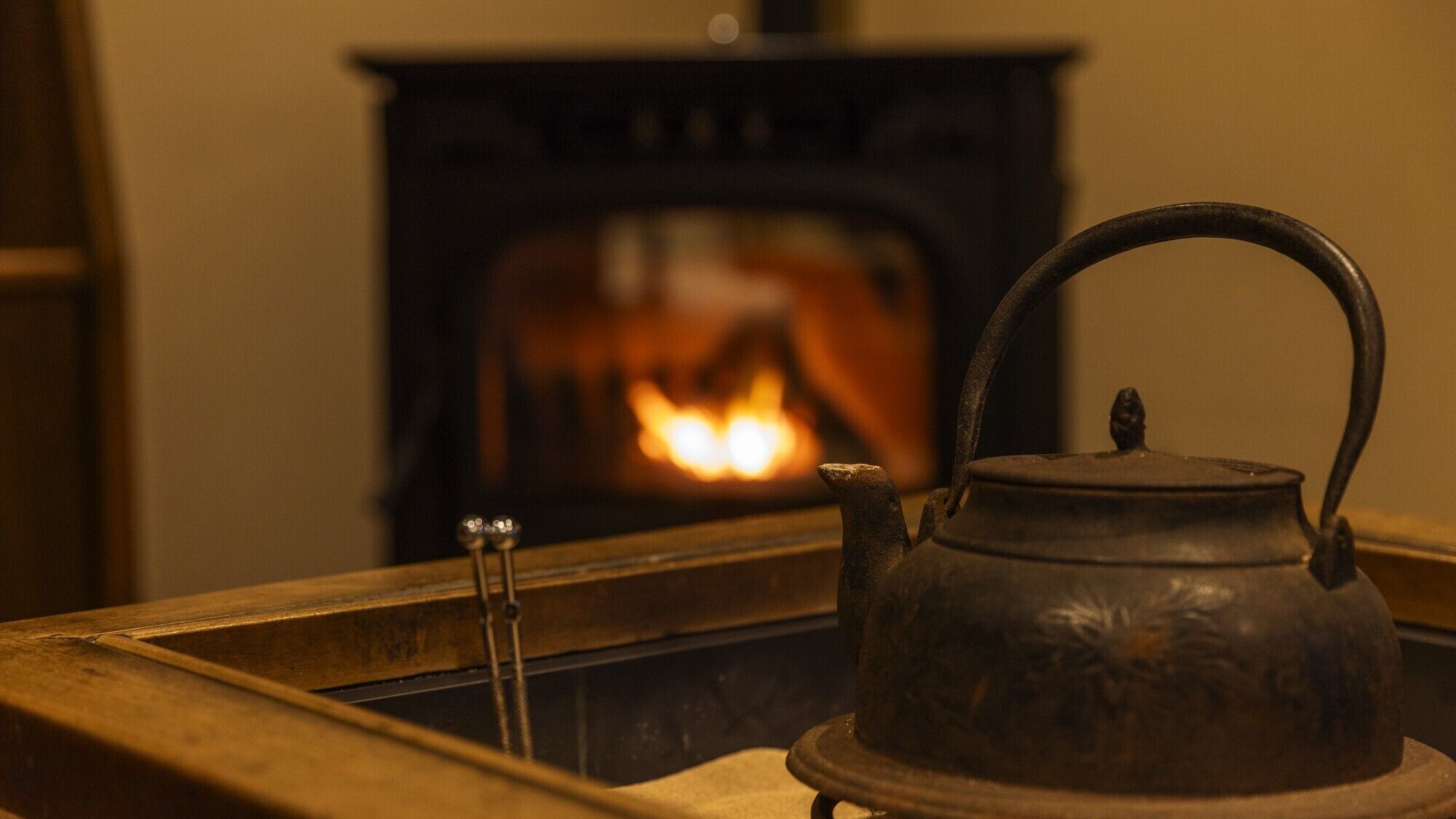 A traditional metal kettle on a wooden tray in front of a fireplace with a visible fire.