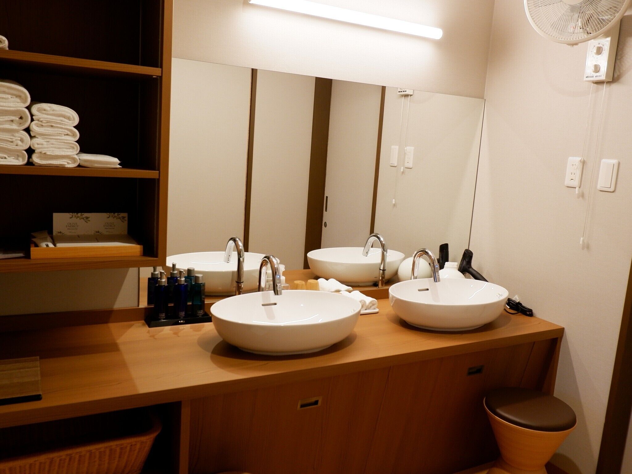 A bathroom with two sinks, a mirror, a fan, and a shelf with towels.