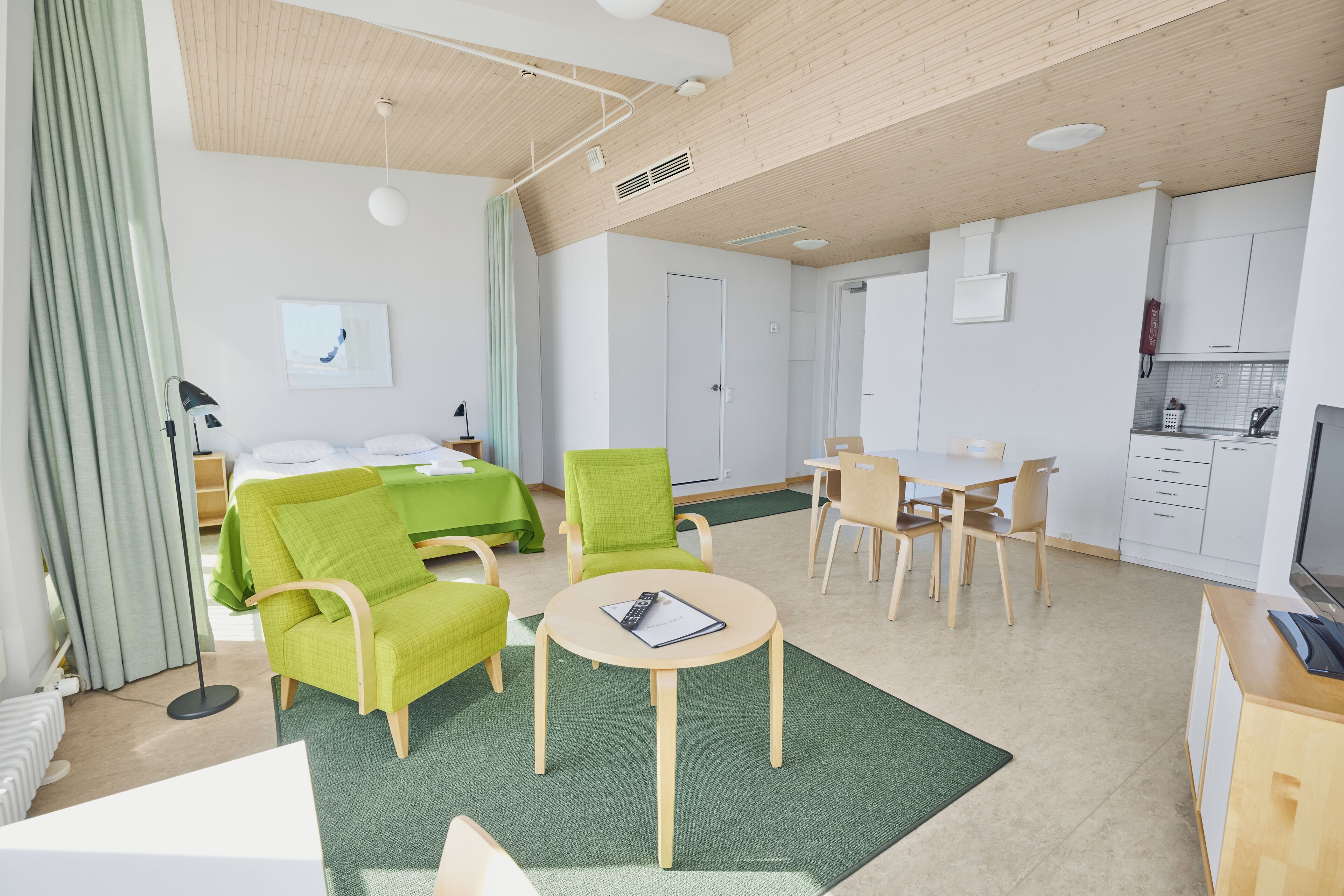 Una habitación de hotel moderna con una cama, sillas verdes, una mesita, un escritorio y una kitchenette.