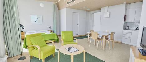 Una habitación de hotel moderna con una cama, sillas verdes, una mesita, un escritorio y una kitchenette.