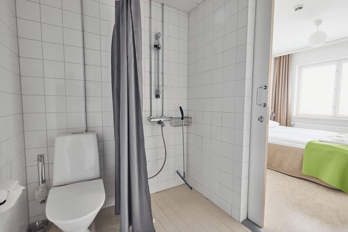 A bathroom with a shower, toilet, and a bed with a green bedspread in the adjacent room.