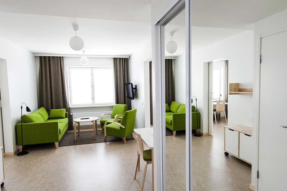 A modern living room with a green sofa, a small table, and a chair. There is a window with curtains, a flat-screen TV mounted on the wall, and a sliding glass door leading to another room.
