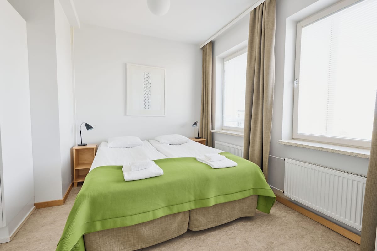 A neatly arranged bedroom with a bed, bedside tables, a radiator, and a window with curtains.