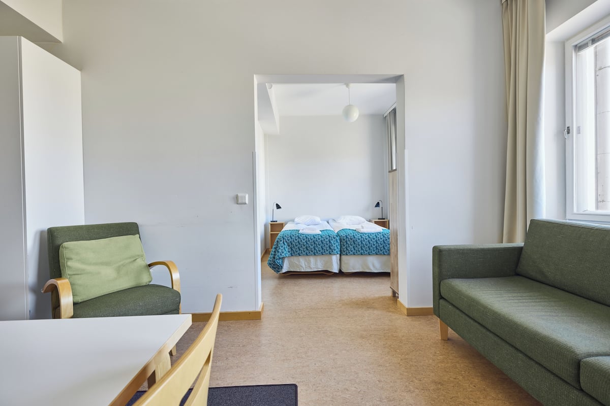 A room with a green sofa, a wooden chair, a small table, and a bed with blue bedding.