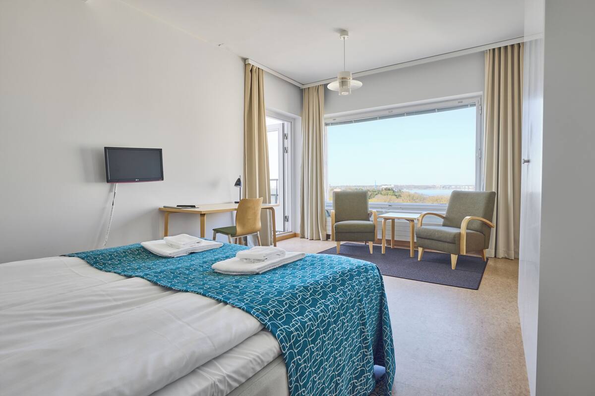 A hotel room with a bed, a desk, a chair, a TV, and a view of the outdoors.